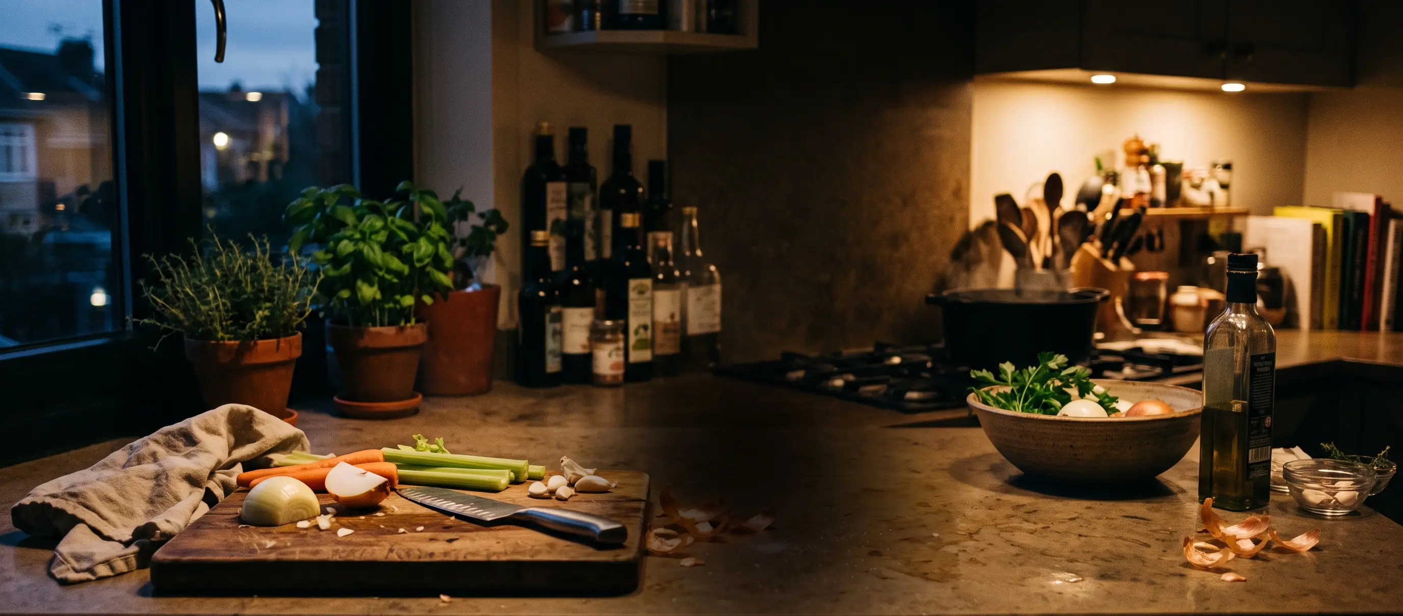 A dim home kitchen at dusk, mid-prep: chopped vegetables on a board, herbs on the sill